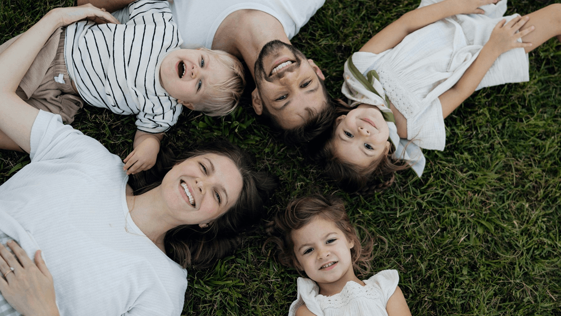 A mom lying on the grass with her three young children, all smiling up at the camera